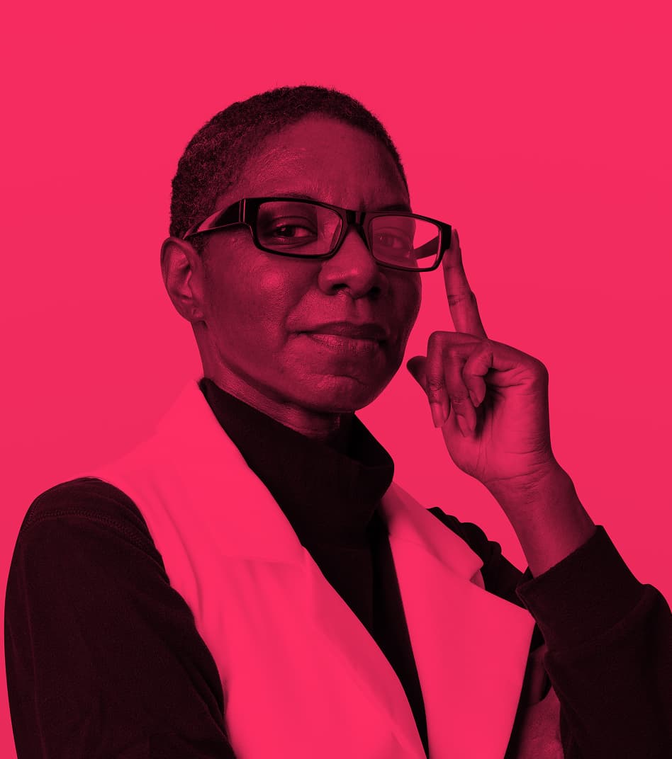 A confident woman with short hair and glasses, wearing a white vest over a dark shirt, touching the side of her glasses and looking at the camera. The image is overlaid with a red color filter.